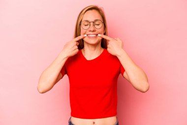 Pembe arka planda izole edilmiş genç beyaz kadın gülümsüyor, parmaklarını ağıza doğrultuyor..