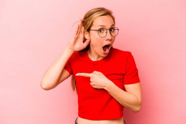 Pembe arka planda izole edilmiş genç beyaz kadın bir dedikoduyu dinlemeye çalışıyor..