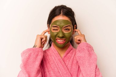 Yüz maskesi takan genç İspanyol kadın beyaz arka planda izole edilmiş kulakları elleriyle kapatıyor..