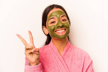 Beyaz arka planda izole edilmiş yüz maskesi takan genç İspanyol kadın neşeli ve kaygısız parmaklarıyla barış sembolü gösteriyor..