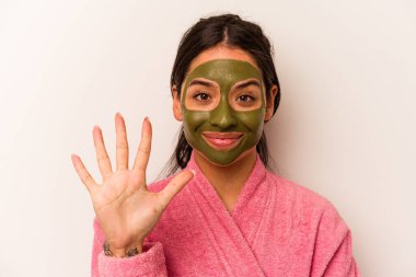 Beyaz arka planda izole edilmiş yüz maskesi takan genç İspanyol kadın gülümseyerek beş numarayı parmaklarıyla gösteriyor..