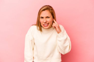Pembe arka planda izole edilmiş genç beyaz bir kadın parmaklarıyla kulaklarını kapatıyor, gürültülü bir ortam yüzünden stresli ve çaresiz..