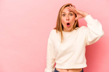 Pembe arka planda izole edilmiş genç beyaz kadın uzaklara bakıyor ve elini alnına koyuyor..