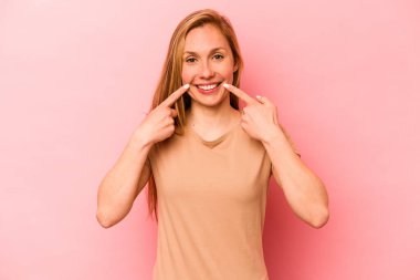Pembe arka planda izole edilmiş genç beyaz kadın gülümsüyor, parmaklarını ağıza doğrultuyor..