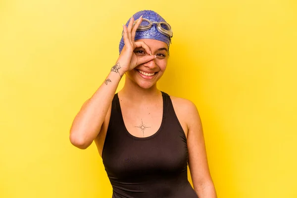 Young swimmer hispanic woman isolated on yellow background excited keeping ok gesture on eye.