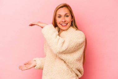 Pembe arka planda izole edilmiş genç beyaz bir kadın işaret parmaklarıyla küçük bir şey tutuyor, gülümsüyor ve kendinden emin..