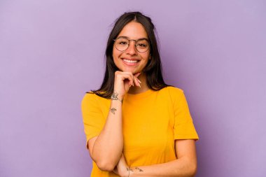 Mor arka planda izole edilmiş genç İspanyol kadın mutlu ve kendinden emin gülümsüyor, elleriyle çenesine dokunuyor..