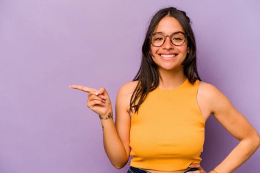 Mor arka planda izole edilmiş genç İspanyol kadın işaret parmağıyla işaret ederek gülümsüyor..