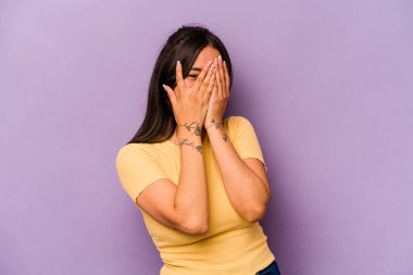 Mor arka planda izole edilmiş genç İspanyol kadın kameraya parmaklarıyla göz kırpıyor, yüzünü kapatmaktan utanıyor..