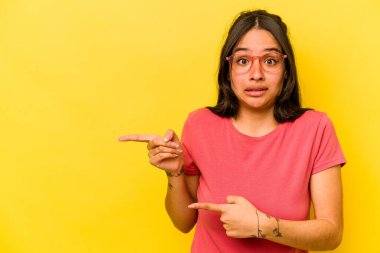 Sarı arka planda izole edilmiş genç İspanyol kadın işaret parmağıyla fotokopi alanını işaret ederek şok oldu..