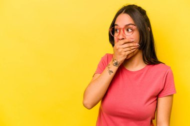 Genç İspanyol kadın sarı arka planda izole edilmiş. Düşünceli bir şekilde bir fotokopi odasına bakıyor. Ağzını eliyle kapatıyor..
