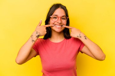 Sarı arka planda izole edilmiş genç İspanyol kadın gülümsüyor, parmaklarını ağzına doğrultuyor..