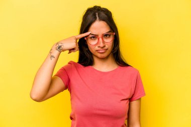 Sarı arka planda izole edilmiş genç İspanyol bir kadın şakağı parmağıyla işaret ediyor, düşünüyor ve bir göreve odaklanıyor..