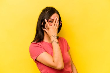 Sarı arka planda izole edilmiş genç İspanyol kadın kameraya parmaklarıyla göz kırpıyor, yüzünü kapatmaktan utanıyor..