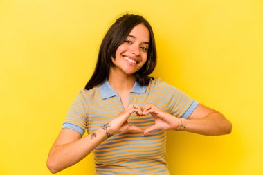 Sarı arka planda izole edilmiş genç İspanyol kadın gülümsüyor ve elleriyle kalp şeklini gösteriyor..