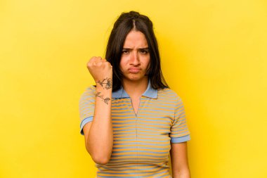 Sarı arka planda izole edilmiş genç İspanyol kadın kameraya yumruk gösteriyor, agresif yüz ifadesi..