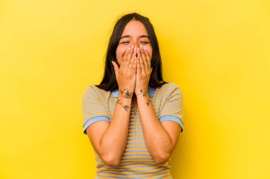 Sarı arka planda izole edilmiş genç İspanyol kadın bir şeye gülüyor, elleriyle ağzını kapatıyor..