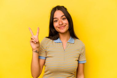 Sarı arka planda izole edilmiş genç İspanyol kadın neşeli ve kaygısız parmaklarıyla barış sembolü gösteriyor..