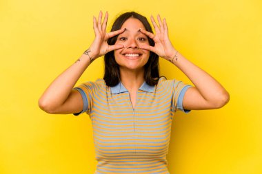 Sarı arka planda izole edilmiş genç İspanyol kadın bir başarı fırsatı bulmak için gözlerini açık tutuyor..