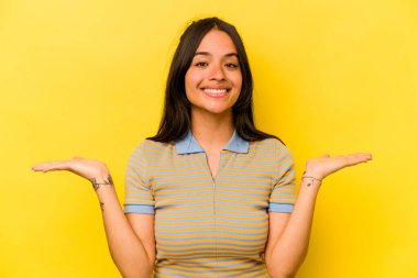 Sarı arka planda izole edilmiş genç İspanyol kadın kollarıyla ölçeği yapıyor, mutlu ve kendinden emin hissediyor..