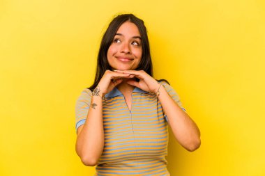 Sarı arka planda izole edilmiş genç İspanyol kadın ellerini çenesinin altında tutuyor ve mutlu bir şekilde yan tarafa bakıyor..
