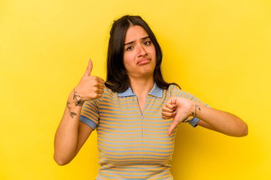 Sarı arka planda izole edilmiş genç İspanyol kadın baş parmağını kaldırıyor, zor bir seçim.