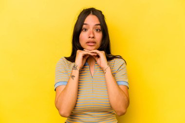 Genç İspanyol kadın sarı arka planda izole edilmiş şans için dua ediyor, şaşırmış ve ağzı açık bir şekilde önüne bakıyor..