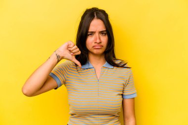 Sarı arka planda izole edilmiş genç İspanyol kadın baş parmağını aşağı gösteriyor, hayal kırıklığı konsepti..