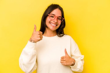 Sarı arka planda izole edilmiş genç İspanyol kadın iki parmağını da kaldırıyor, gülümsüyor ve kendinden emin..