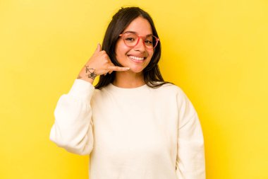 Sarı arka planda izole edilmiş genç İspanyol kadın parmaklarıyla bir cep telefonu hareketi gösteriyor..