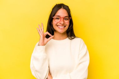 Sarı arka planda izole edilmiş genç İspanyol kadın bir göz kırptı ve elleriyle iyi bir jest yaptı..