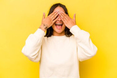 Sarı arka planda izole edilmiş genç İspanyol kadın elleriyle gözlerini kapatıyor, geniş bir gülümsemeyle bir sürpriz bekliyor..