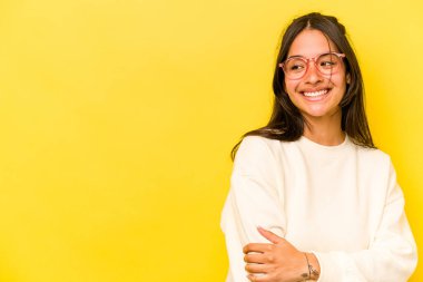 Sarı arka planda izole edilmiş genç İspanyol kadın kollarını kavuşturarak gülümsüyor..