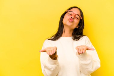 Sarı arka planda izole edilmiş genç İspanyol kadın dudakları katlıyor ve hava öpücüğü göndermek için avuç içleri tutuyor..