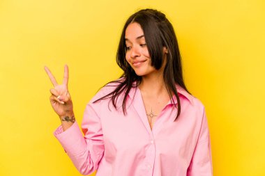 Sarı arka planda izole edilmiş genç İspanyol kadın neşeli ve kaygısız parmaklarıyla barış sembolü gösteriyor..