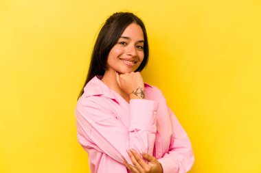 Sarı arka planda izole edilmiş genç İspanyol kadın mutlu ve kendinden emin gülümsüyor, elleriyle çenesine dokunuyor..