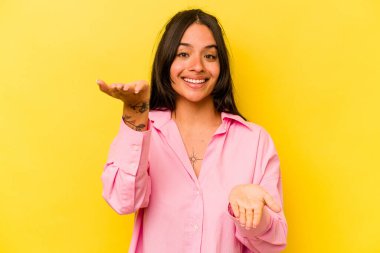 Sarı arka planda izole edilmiş genç İspanyol kadın kollarıyla ölçeği yapıyor, mutlu ve kendinden emin hissediyor..