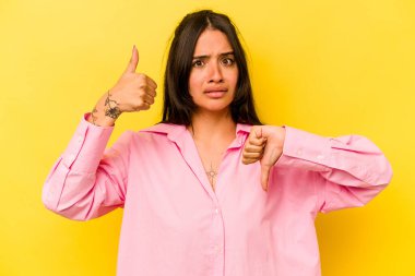 Sarı arka planda izole edilmiş genç İspanyol kadın baş parmağını kaldırıyor, zor bir seçim.