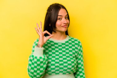 Sarı arka planda izole edilmiş genç İspanyol kadın bir göz kırptı ve elleriyle iyi bir jest yaptı..