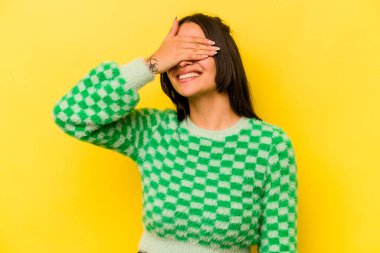 Sarı arka planda izole edilmiş genç İspanyol kadın elleriyle gözlerini kapatıyor, geniş bir gülümsemeyle bir sürpriz bekliyor..