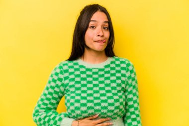 Sarı arka planda izole edilmiş genç İspanyol kadın karnına dokunuyor, nazikçe gülümsüyor, yemek yiyor ve tatmin oluyor..