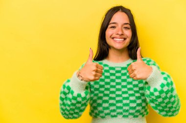 Sarı arka planda izole edilmiş genç İspanyol kadın gülümsüyor ve başparmağını kaldırıyor.