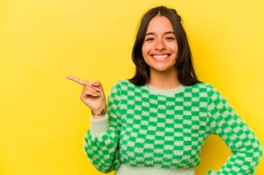 Sarı arka planda izole edilmiş genç İspanyol kadın işaret parmağıyla işaret ederek gülümsüyor..