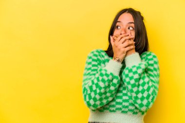 Genç İspanyol kadın sarı arka planda izole edilmiş. Düşünceli bir şekilde bir fotokopi odasına bakıyor. Ağzını eliyle kapatıyor..