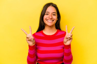 Sarı arka planda izole edilmiş genç İspanyol kadın zafer işareti gösteriyor ve gülümsüyor..