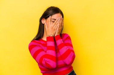 Sarı arka planda izole edilmiş genç İspanyol kadın kameraya parmaklarıyla göz kırpıyor, yüzünü kapatmaktan utanıyor..