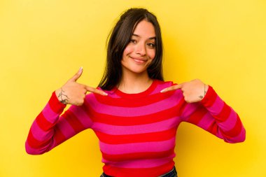 Sarı arka planda izole edilmiş genç İspanyol kadın gömlek fotokopi alanını elle işaret ediyor, gururlu ve kendinden emin.