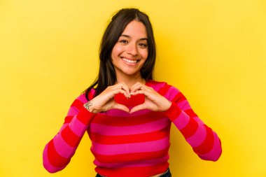 Sarı arka planda izole edilmiş genç İspanyol kadın gülümsüyor ve elleriyle kalp şeklini gösteriyor..