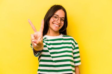 Sarı arka planda izole edilmiş genç İspanyol kadın neşeli ve kaygısız parmaklarıyla barış sembolü gösteriyor..