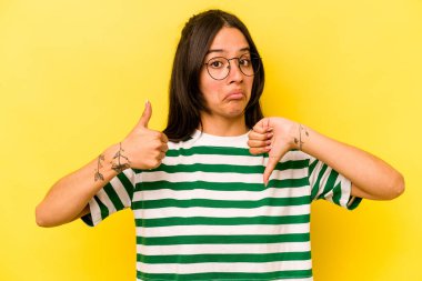 Sarı arka planda izole edilmiş genç İspanyol kadın baş parmağını kaldırıyor, zor bir seçim.